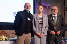Drei formal gekleidete Personen stehen nebeneinander und lächeln in die Kamera. Der Mann auf der rechten Seite trägt einen Anzug mit einer feierlichen Kette. Im Vordergrund steht ein Tisch mit einem Blumenarrangement, und hinter ihnen befindet sich eine Präsentationswand.