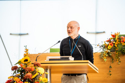 Ein Mann steht an einem Podium mit Mikrofonen, trägt einen schwarzen Pullover und eine beigefarbene Hose und spricht in einem hell erleuchteten, mit bunten Blumenarrangements geschmückten Raum.