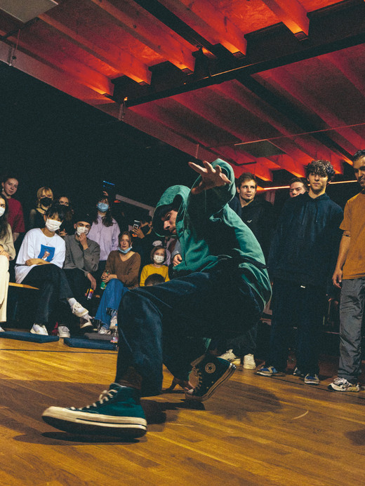 A dancer in a teal hoodie and sneakers performs a dynamic move on a wooden floor in front of an audience. The crowd, some wearing masks, watches attentively in a dimly lit room with red ceiling lights.