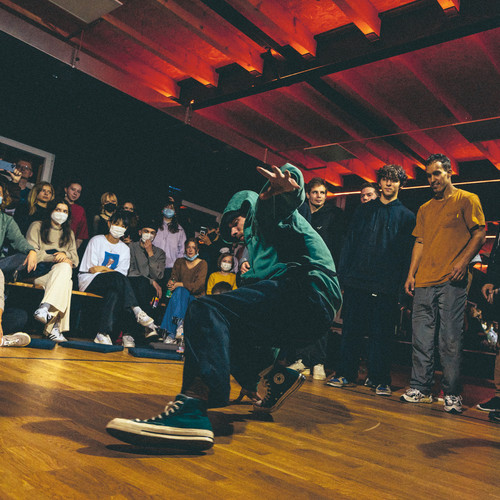 A dancer in a teal hoodie and sneakers performs a dynamic move on a wooden floor in front of an audience. The crowd, some wearing masks, watches attentively in a dimly lit room with red ceiling lights.