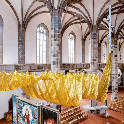 Innenansicht einer Kirche mit hohen Bogenfenstern, Steinsäulen und einer großen, von der Decke hängenden Installation aus goldenem Stoff. Darunter sind Reihen von Holzstühlen angeordnet, und links ist ein dekoriertes Paneel zu sehen.