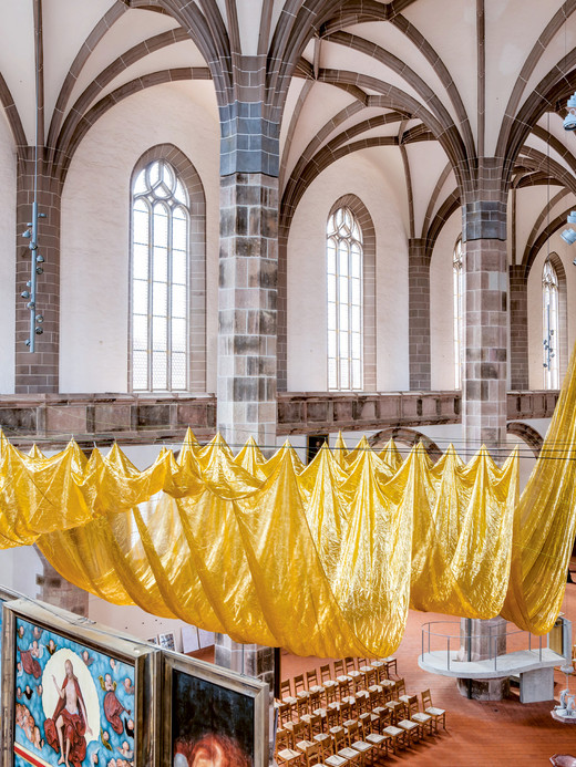 Innenansicht einer Kirche mit hohen Bogenfenstern, Steinsäulen und einer großen, von der Decke hängenden Installation aus goldenem Stoff. Darunter sind Reihen von Holzstühlen angeordnet, und links ist ein dekoriertes Paneel zu sehen.