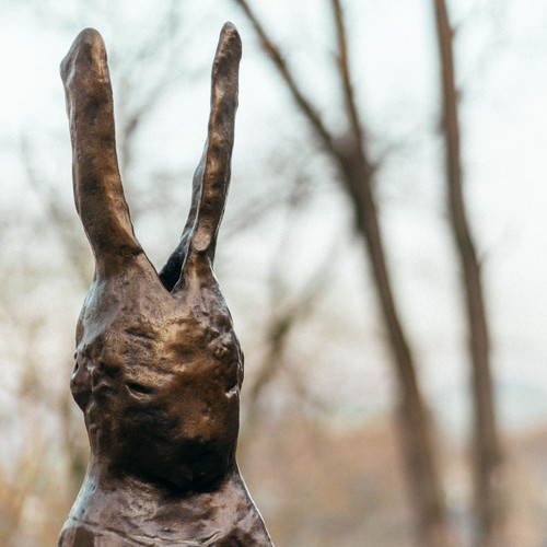 Eine Bronzeskulptur eines Kaninchens mit langen Ohren steht im Freien, umgeben von verschwommenen Bäumen und einem weichen, gedämpften Hintergrund.