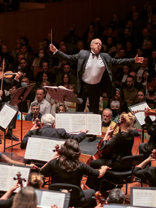 Ein Dirigent leitet leidenschaftlich ein Orchester, dessen Musiker in Reihen sitzen und ihre Instrumente spielen. Im Hintergrund ist das Publikum zu sehen, das gespannt der Aufführung in einem Konzertsaal zusieht.