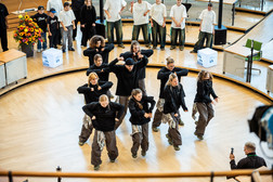 Eine Gruppe von Tänzern in schwarzer Kleidung tritt in Formation auf einem Holzboden auf, während Personen in weißen Hemden von hinten zusehen. Eine Person filmt die Szene, und links ist ein Blumengesteck zu sehen.