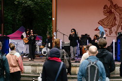 A band plays on an outdoor stage while people in jackets and hoodies look on. The background of the stage shows an illustration of dancing figures, and trees can be seen in the background.