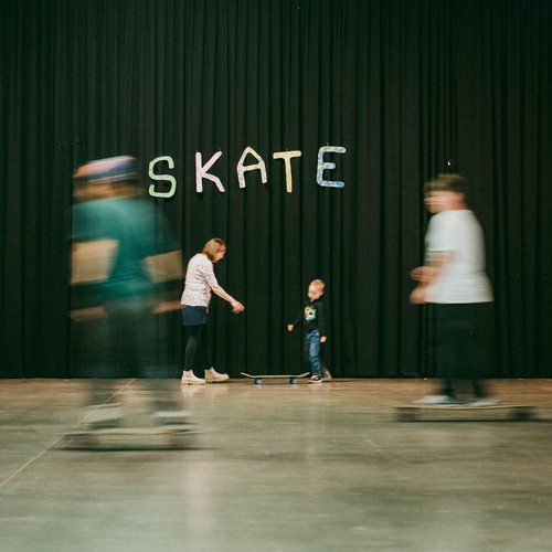 Ein Kind steht mit einem Erwachsenen vor einem schwarzen Vorhang, über dem das Wort "SKATE" steht. Mehrere unscharfe Personen auf Skateboards bewegen sich um sie herum auf einem glatten Hallenboden.