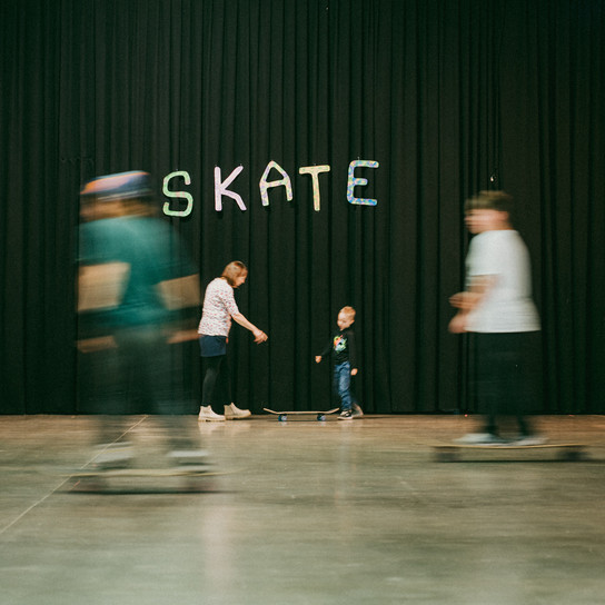 Ein Kind steht mit einem Erwachsenen vor einem schwarzen Vorhang, über dem das Wort "SKATE" steht. Mehrere unscharfe Personen auf Skateboards bewegen sich um sie herum auf einem glatten Hallenboden.