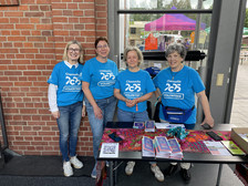 Vier Frauen in passenden blauen "Chemnitz Volunteer"-Shirts stehen lächelnd hinter einem Informationstisch mit Broschüren, Flyern und bunten Tüchern in einem Gebäude mit Backsteinwänden und großen Fenstern.