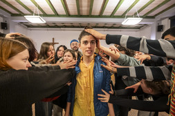 Eine Person steht in der Mitte eines Raumes und blickt mit ernster Miene nach vorne, während viele Menschen um sie herum ihre Hände ausstrecken, um ihren Kopf und ihre Schultern zu berühren. Im Hintergrund ist ein Holzkreuz zu sehen.