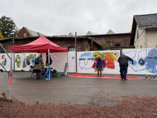 On a wet, cloudy day, people paint colourful graffiti artwork on a white barricade under a red tent roof, with brick buildings in the background.