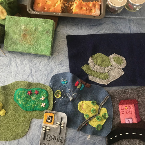 A table is covered with felt crafts, including a tree, a pond, buildings, and a road. Nearby are green yarn, a green sponge, and a tray of baked goods with partially consumed squares. Two small red jars are visible in the corner.