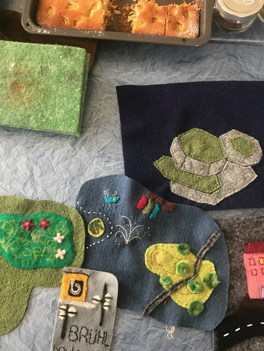 A table is covered with felt crafts, including a tree, a pond, buildings, and a road. Nearby are green yarn, a green sponge, and a tray of baked goods with partially consumed squares. Two small red jars are visible in the corner.