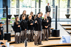 A group of performers in matching black shirts and grey trousers stand together in an interior space. One person in the foreground is holding a microphone and gesticulating, while others stand behind, some smiling. In the background are large windows and a seated audience.