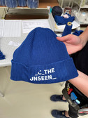 Eine Person hält eine blaue Strickmütze mit den Worten „THE UNSEEN“ in Weiß gestickt. Die Mütze hat eine gerippte Textur. Die Person steht in einem Arbeitsbereich, im Hintergrund sind verschiedene Werkzeuge und blaue Garnkegel zu sehen.