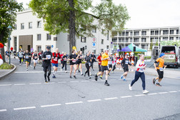 A group of runners in sportswear take part in a road race on a city street. Buildings, trees and a tent can be seen in the background. Spectators watch from the pavement.