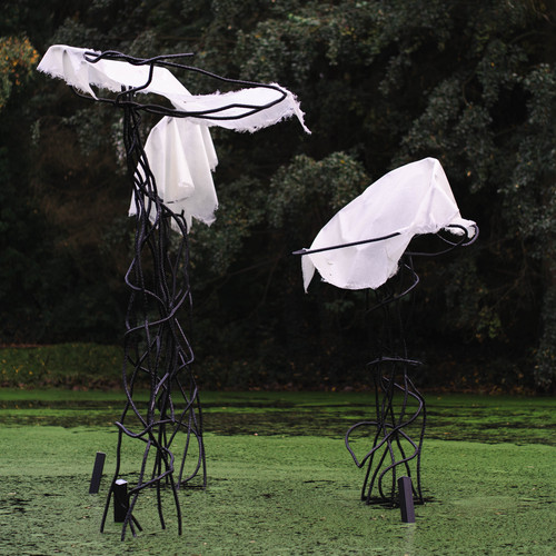 Zwei hohe, abstrakte schwarze Metallskulpturen stehen in einem grünen, mit Algen bewachsenen Teich, der mit weißem Stoff drapiert ist, und im Hintergrund stehen dichte Bäume.