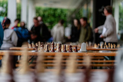 Several wooden chess boards are set up on long tables outside, ready to be played. The chess pieces are set up and a digital timer is visible. In the background, people are gathered and blurred, indicating an event.