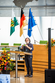 Ein Mann mit grauen Haaren und Bart steht an einem Podium und hält einen Vortrag in einer modernen Halle. Im Hintergrund sind bunte Blumengestecke und mehrere Flaggen zu sehen, darunter die deutsche und die der Europäischen Union.