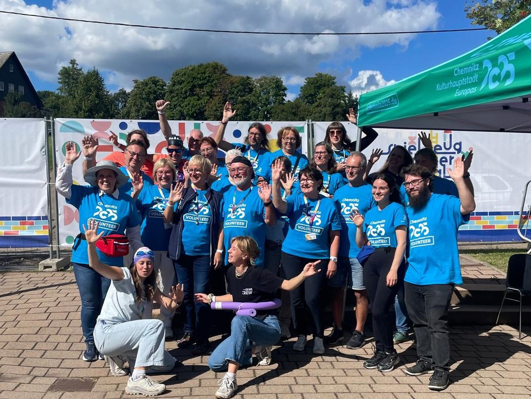 A group of people in matching blue shirts pose and wave under a sunny sky at an outdoor event. They are standing near a tent with the word "25" and colourful decorations in the background.