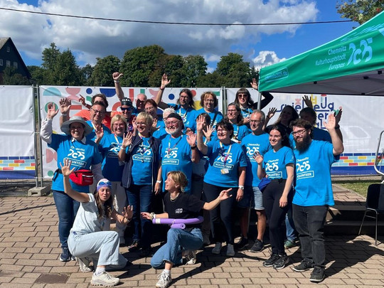 A group of people in matching blue shirts pose and wave under a sunny sky at an outdoor event. They are standing near a tent with the word "25" and colourful decorations in the background.