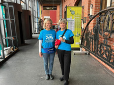 Zwei Frauen in blauen "Chemnitz 2025 Volunteer"-Shirts stehen nebeneinander in einem modernen Gebäude mit Backsteinwänden und großen Fenstern. Beide lächeln in die Kamera, eine hält einen roten Gegenstand, im Hintergrund sind Veranstaltungsbanner zu sehen.