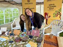 Zwei lächelnde Frauen stehen hinter einem Tisch, auf dem verschiedene Schokoladenprodukte an einem Marktstand im Freien angeboten werden. Auf dem Tisch liegen beschriftete Schokoladentafeln und Proben. Hinter ihnen ist ein gelbes Transparent über Kakao zu sehen.