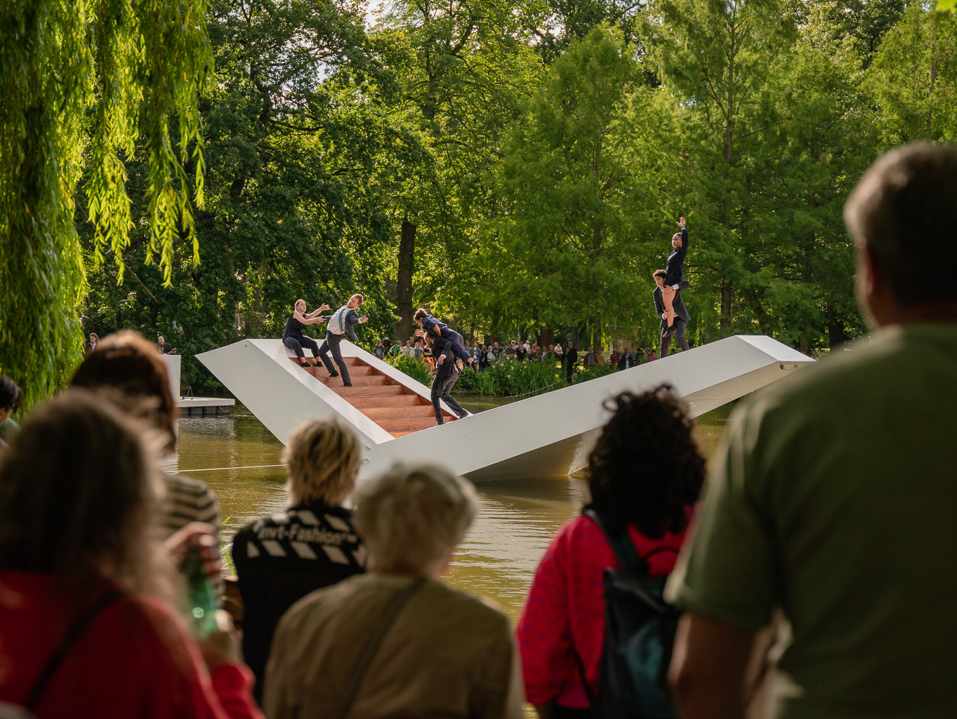 A large crowd gathers in a lush green park, watching a group of performers on a tilted white platform over a pond. The platform appears to be balancing precariously. Some trees and clouds are visible in the background.