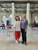 Two women stand together smiling in a bright room with colourful chairs, high windows and high ceilings. One is wearing a white shirt and red trousers, the other a striped shirt and a headband. People are sitting in the background.