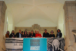 A group of twelve people stands behind a blue sign reading “Edvard Munch Angst,” featuring artwork from “The Scream,” in a grand stone building with arches and decorative details above them.