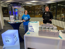 Hinter einem Promotiontisch mit Broschüren stehen zwei Frauen. Sie tragen blaue Shirts mit der Aufschrift „Chemnitz Kulturhauptstadt Europas 2025“. Zum Aufbau gehören beleuchtete Würfel mit dem Text „C the Unseen“.