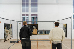 Two people stand in front of fenced-in art displays and look at paintings in a bright, modern gallery space with white walls, high windows and artworks depicting people in muted colours.
