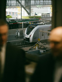 Blick auf einen modernen Zug auf einer Eisenbahnstrecke mit verschwommenen Silhouetten von zwei Personen im Vordergrund. Die Szene spielt in einem Bahnhof mit Industrieelementen und einem weiteren im Hintergrund sichtbaren Fahrzeug.