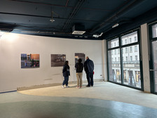 Three people stand in front of three works of art displayed on a white wall in a brightly lit gallery space with large windows overlooking a city street.