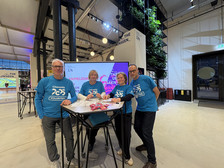 Vier Freiwillige in passenden blauen "Chemnitz 2025"-Shirts stehen hinter einem Tisch mit Papieren und rosafarbenen Schlüsselbändern und lächeln in die Kamera in einem geräumigen, modernen Saal mit hohen Decken und Grünpflanzen.