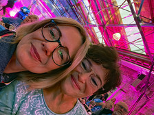Two women smile close together in a lively, pink-lit interior with a glass ceiling. Other people are seated in the background, suggesting a social gathering or event.