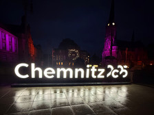 Großes beleuchtetes "Chemnitz 2025"-Schild bei Nacht auf einem Stadtplatz, mit historischen Gebäuden und einer Kirche im Hintergrund, die beide mit lila Lichtern beleuchtet sind. Das nasse Pflaster reflektiert das Leuchten des Schildes.