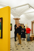 A group of people look at and discuss works of art in a brightly lit art gallery with white walls and vaulted ceilings. A man in the foreground gestures while holding a drink in his hand. Paintings hang on the walls and a striking yellow room divider stands nearby.