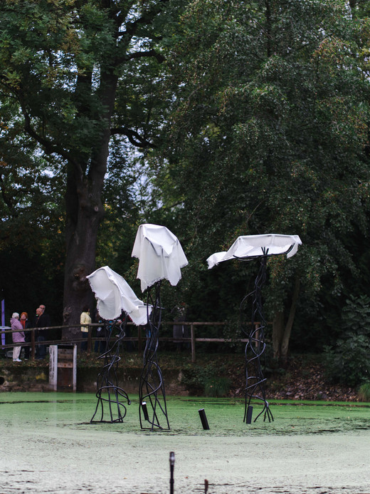 Aus einem mit Grünalgen bewachsenen Teich ragen drei abstrakte Metallskulpturen, die jeweils mit einem weißen Tuch bedeckt sind. Umgeben von Bäumen stehen mehrere Menschen an einem Holzzaun im Hintergrund und betrachten das Kunstwerk.