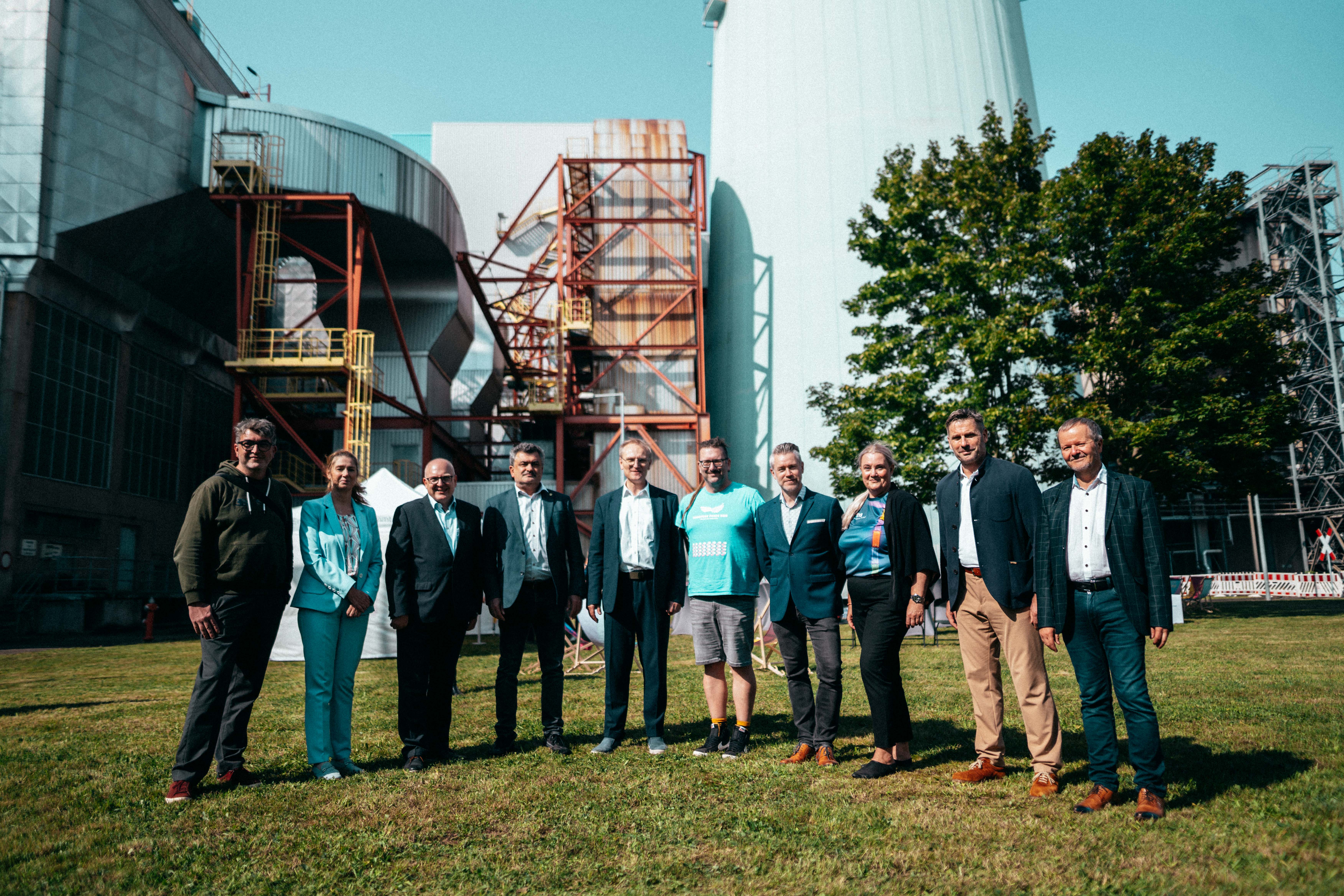 A group of ten people are standing outside on a lawn in front of an industrial backdrop. The group consists of men and women in business and leisure wear. They pose in a row and smile at the camera, with a sunny sky and trees in the background.