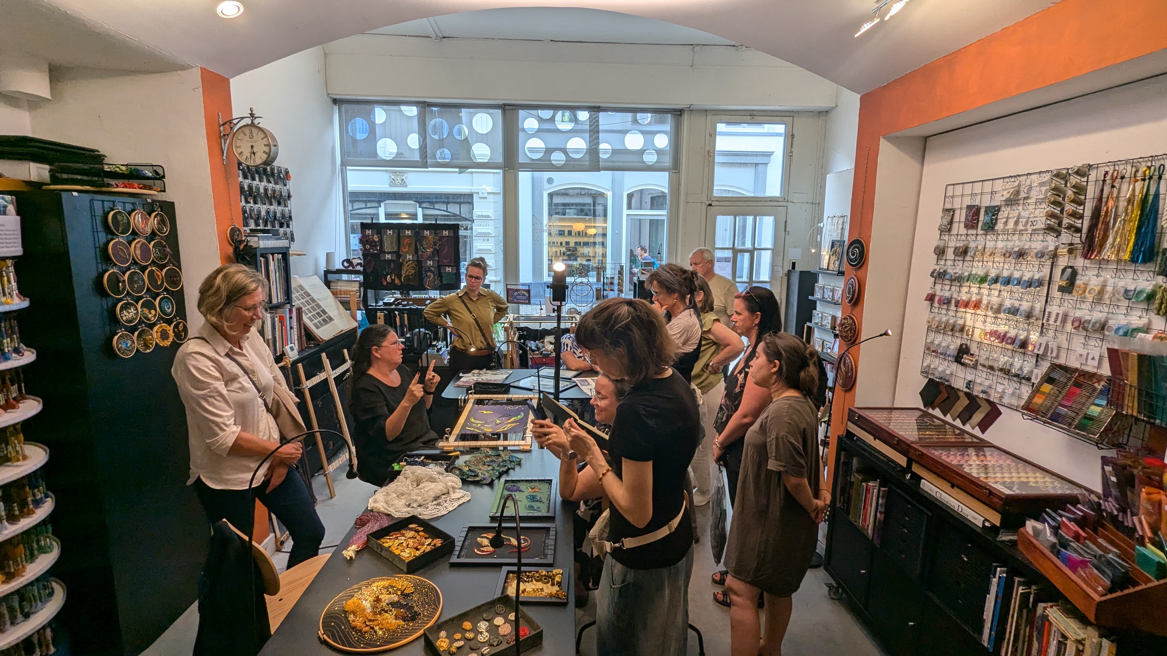 A group of people are gathered in an art shop. Some are standing, while others are seated around a table filled with craft materials. Shelves filled with art supplies line the walls. The atmosphere appears collaborative and focused.