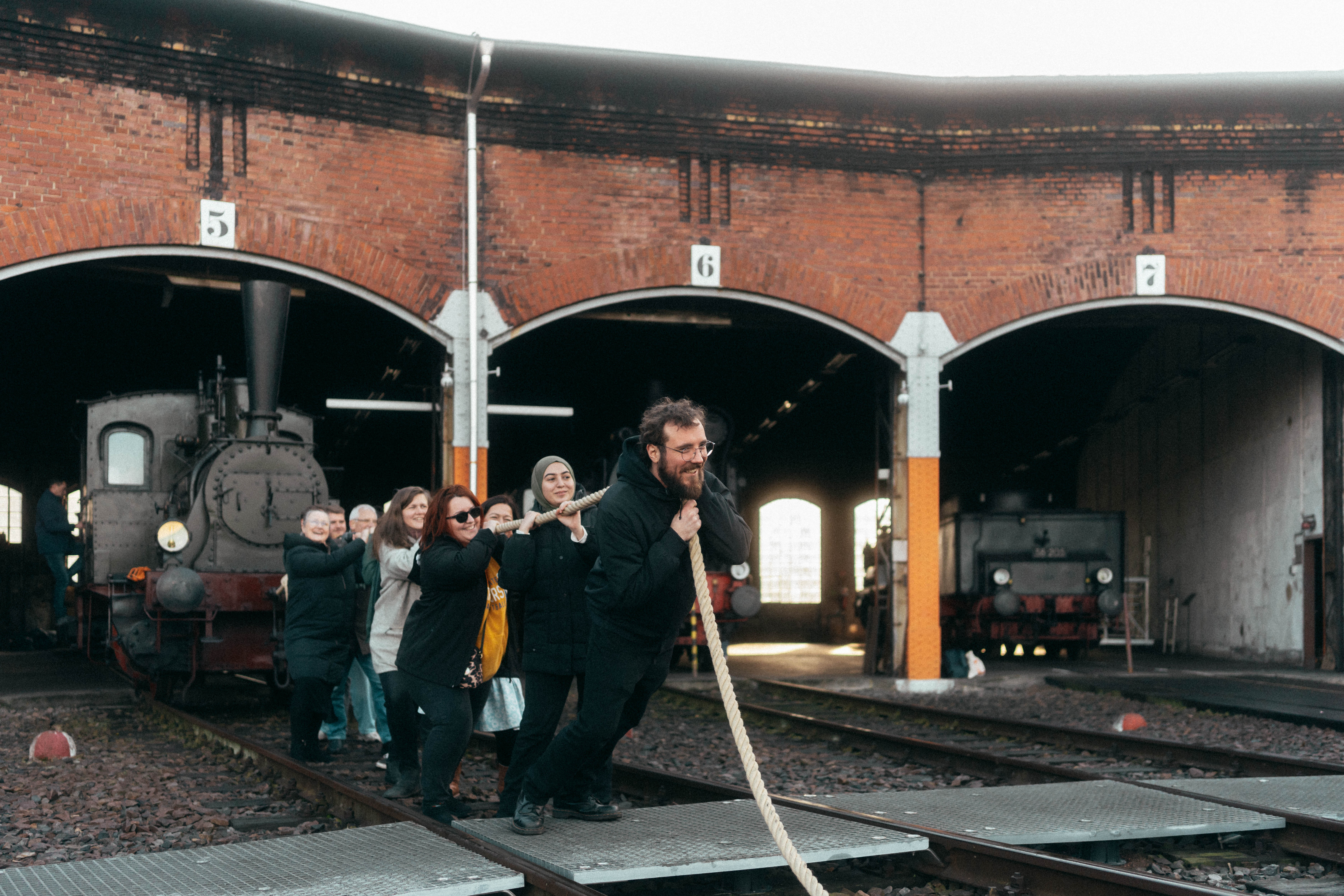 A group of people playfully pull an old steam locomotive out of an engine shed using a thick rope. The engine shed has several open compartments and the scene conveys a fun, communal effort.