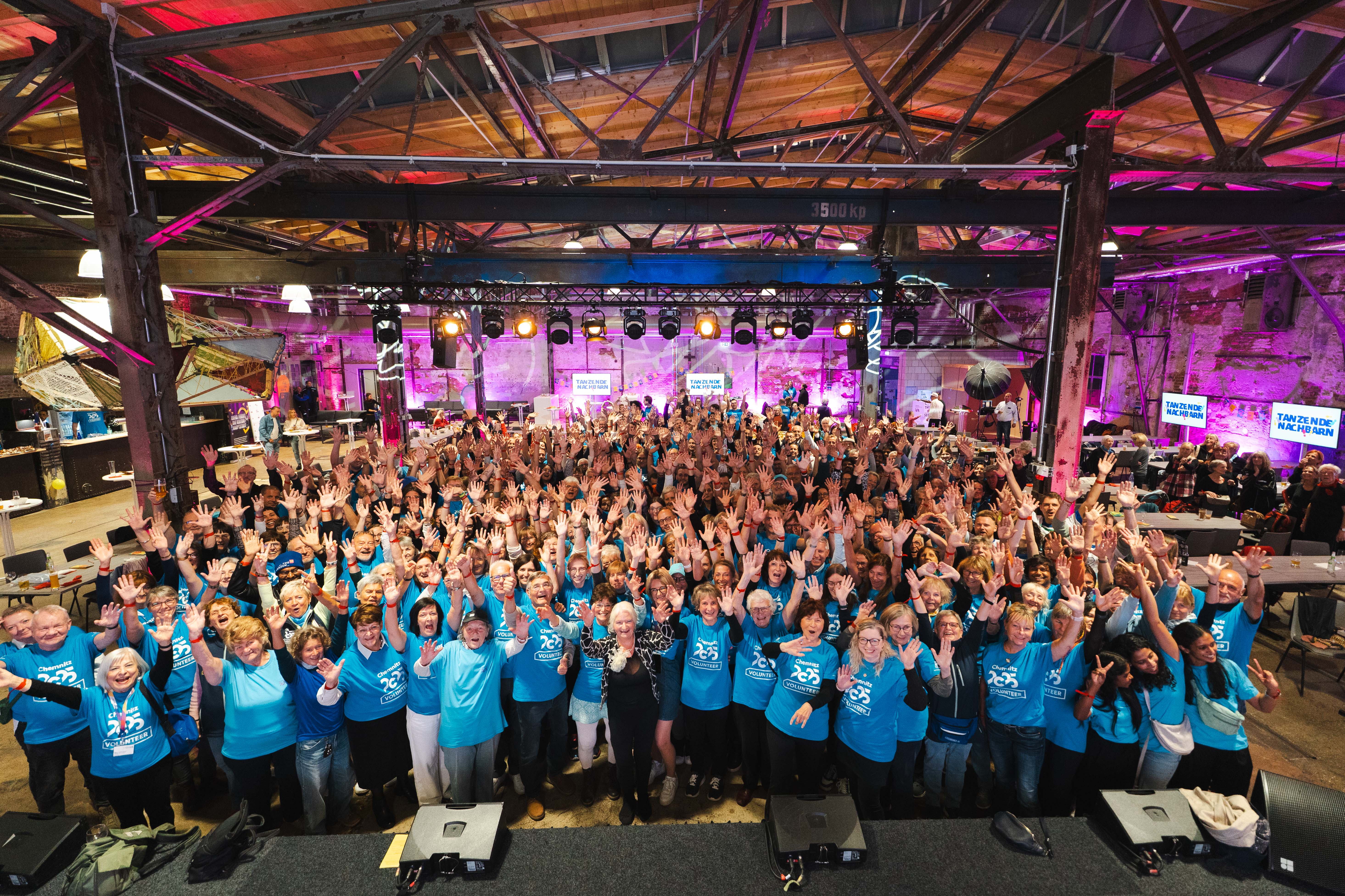 A large group of people in matching blue shirts stand together in a spacious, industrial-like room with exposed beams and colourful stage lights, smiling and waving at the camera.