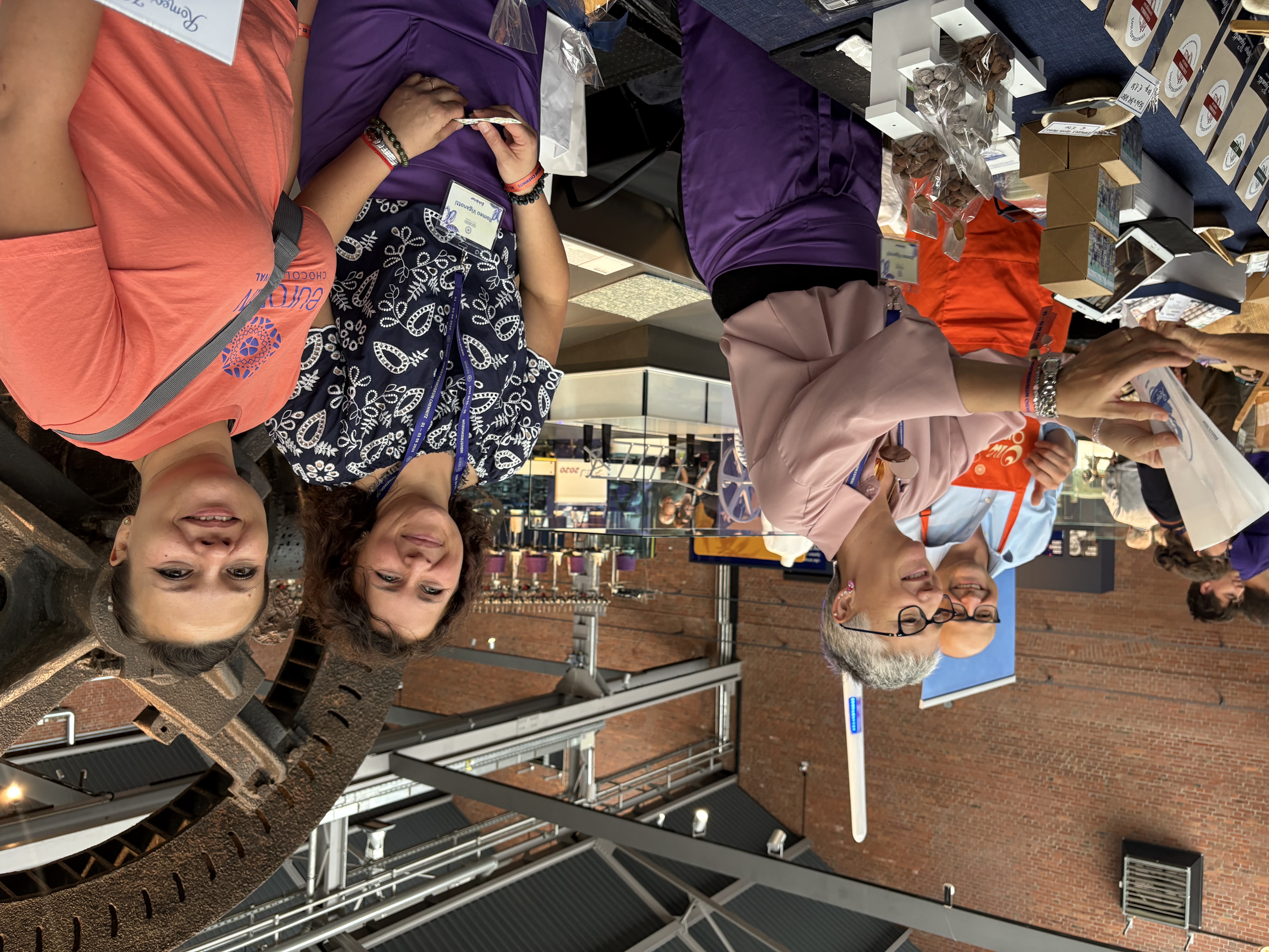 Four smiling people stand behind a table with various chocolates and treats at a market or event in a hall, with industrial machinery and brick walls in the background. A woman hands an item to a customer.