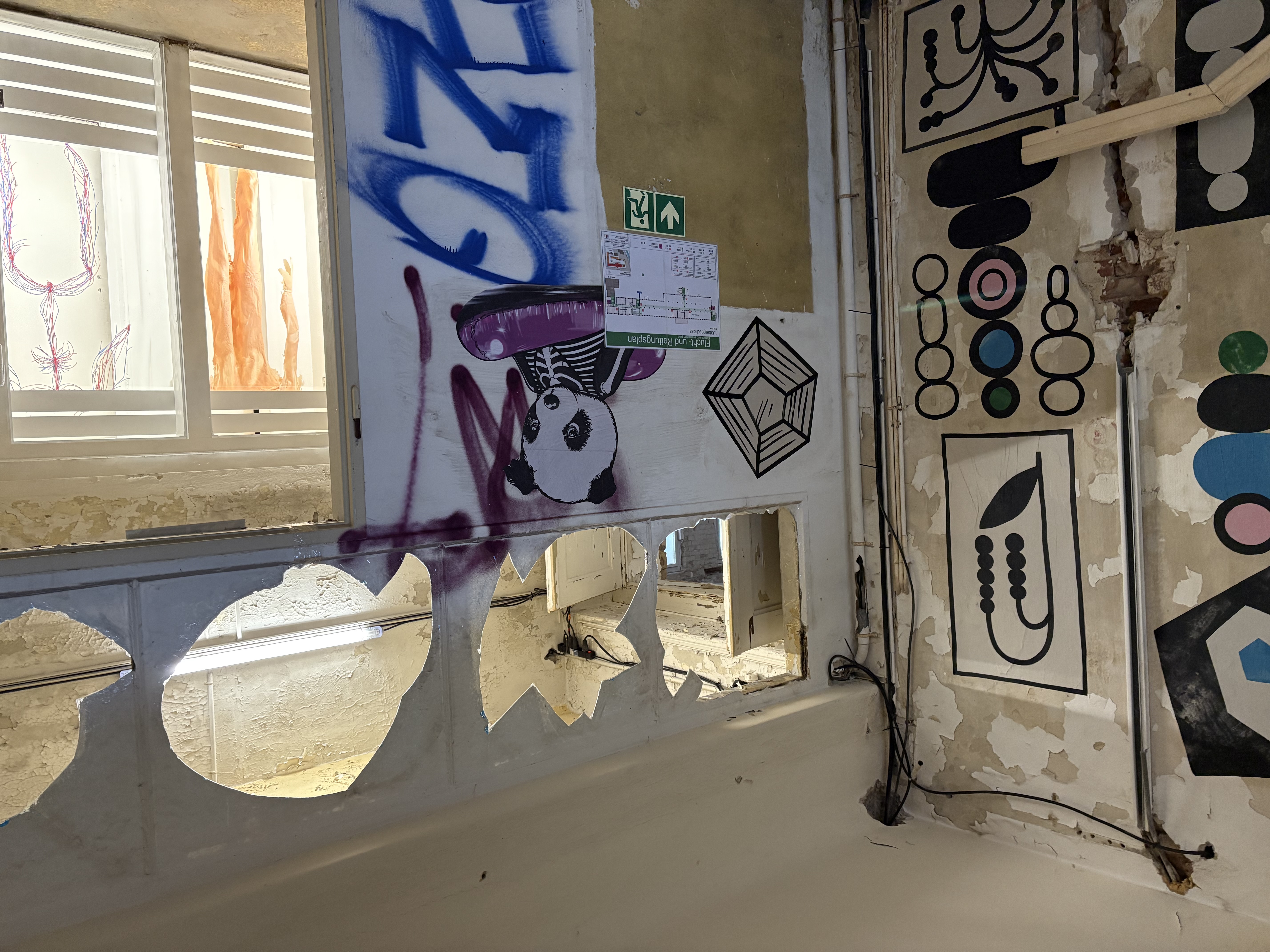 A room with peeling paint, geometric wall design and graffiti with a panda on a purple bumper car. There are exposed wires, emergency exit signs and a window with cut-out arches above the door.