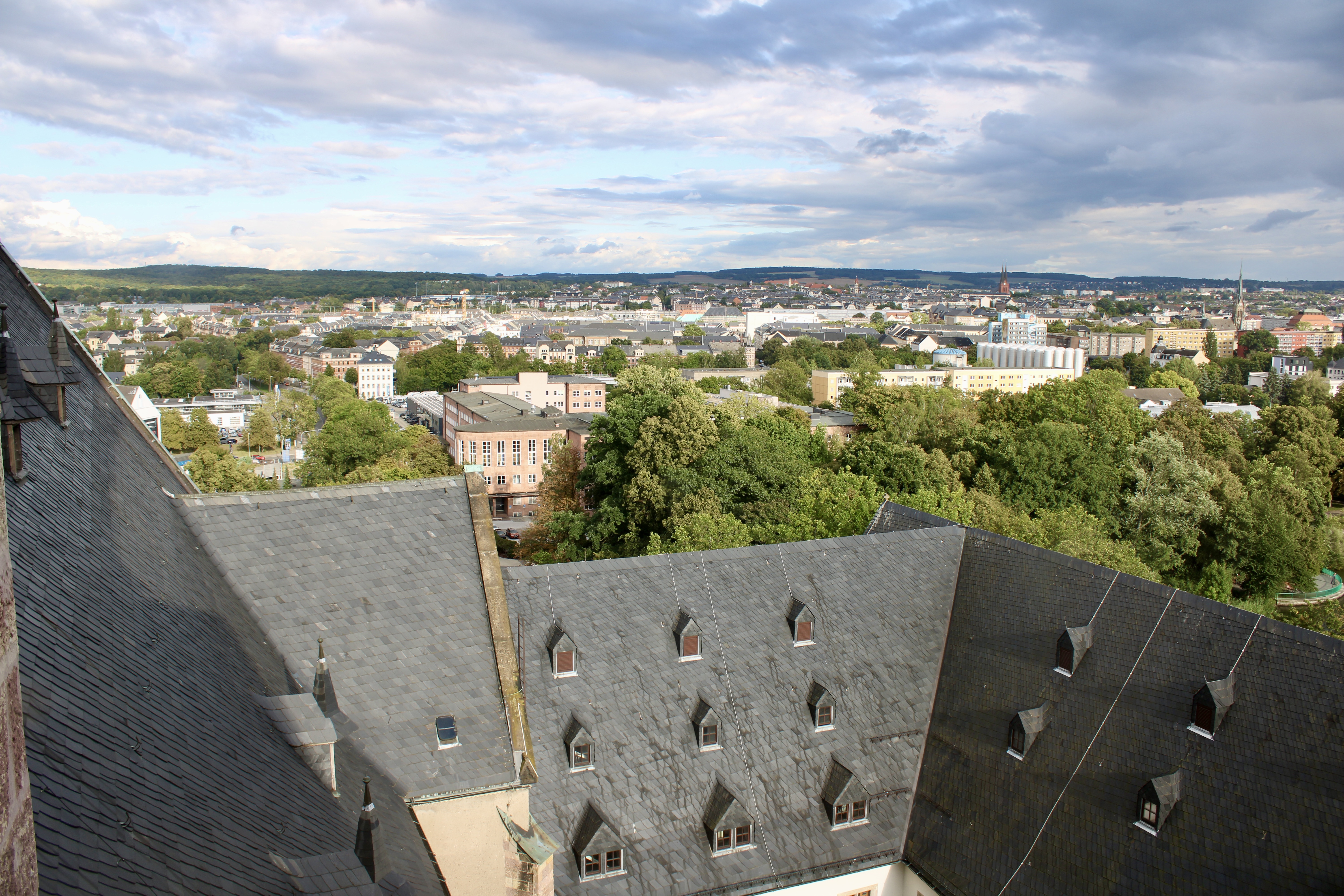 A view of the city from above shows slate-covered roofs with triangular windows, green trees and a sprawling city stretching into the distance under a partly cloudy sky.