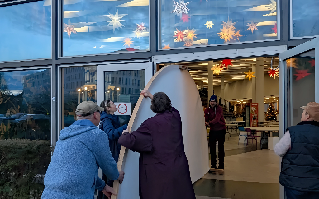 Four people work together to bring a large, curved object through double glass doors into a brightly lit building decorated with colourful star-shaped lights and a Christmas tree inside.