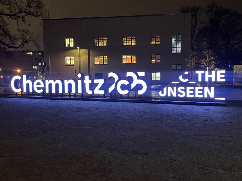 Illuminated sign with the inscription "Chemnitz 2025 - C THE UNSEEN" against a night-time backdrop. The sign is brightly lit and thus contrasts with the dark surroundings. A building and leafless trees can be seen behind it.