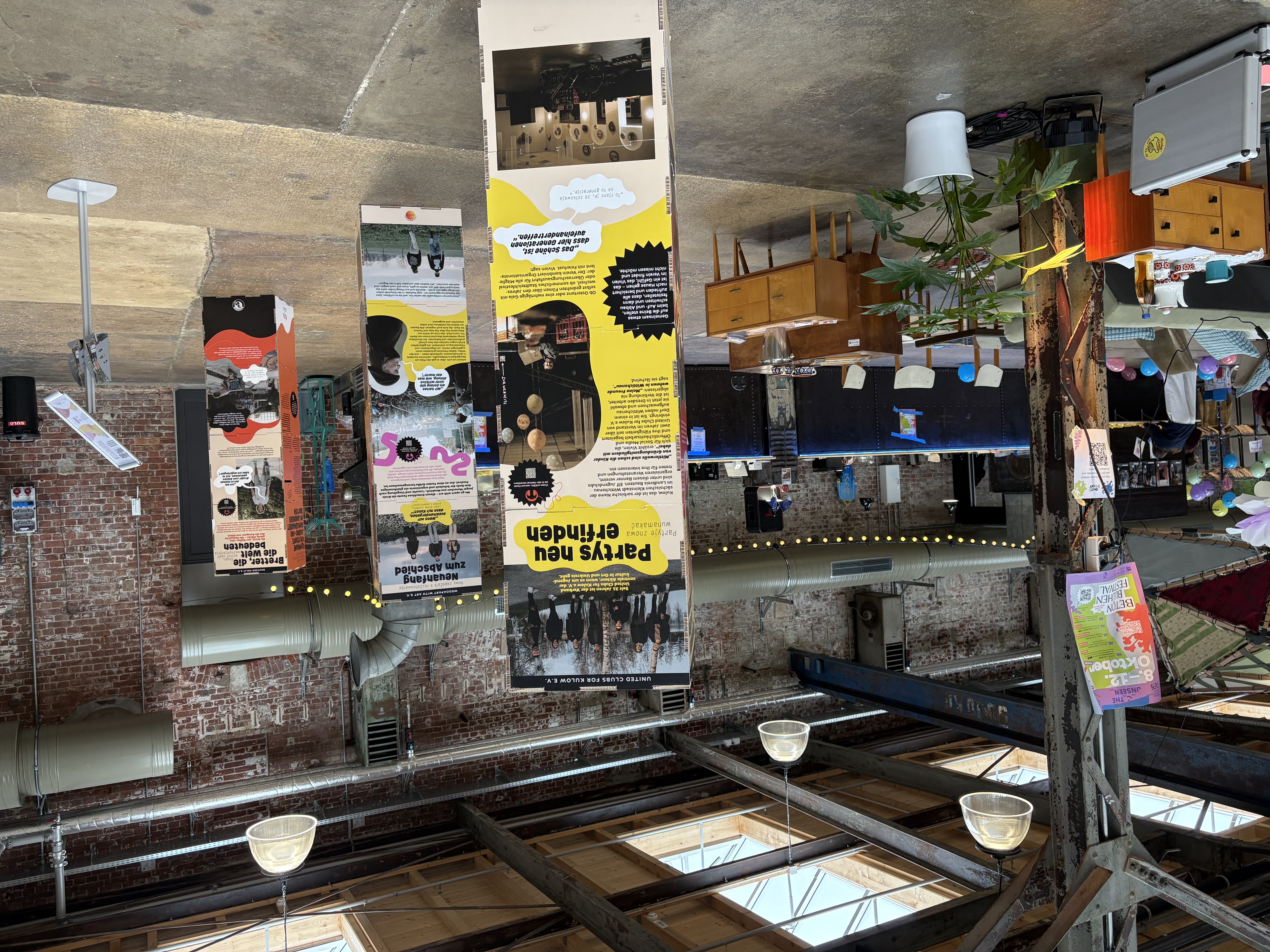 A covered event space with exposed brick walls and industrial beams features information boards, retro furniture, potted plants and people in the background. Sunlight shines through the skylights on the ceiling.