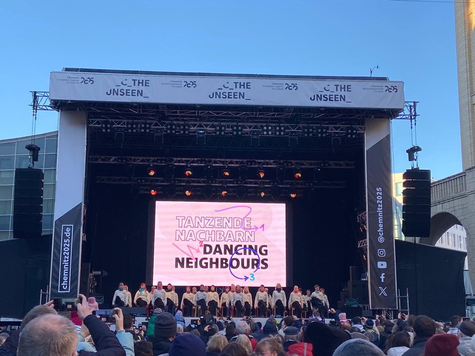 A large crowd watches a group of dancers performing on an open-air stage. The text "Dancing neighbours" can be read on the screen behind them. The event appears to be part of "Chemnitz 2025".